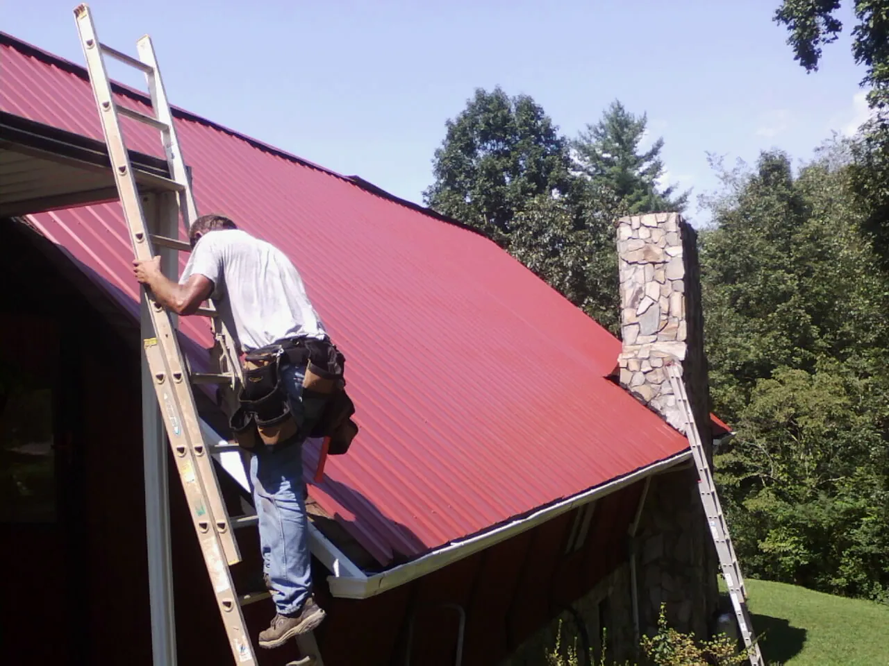 Roofer on a ladder beside a red metal roof and stone chimney — Soffit & Fascia Repair by Roofing Brothers in La Pine